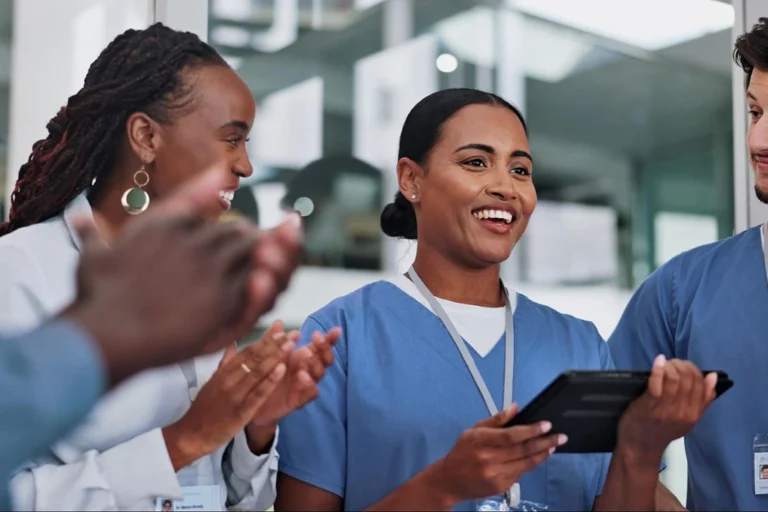 Healthcare workers in scrubs smiling and using a tablet together.